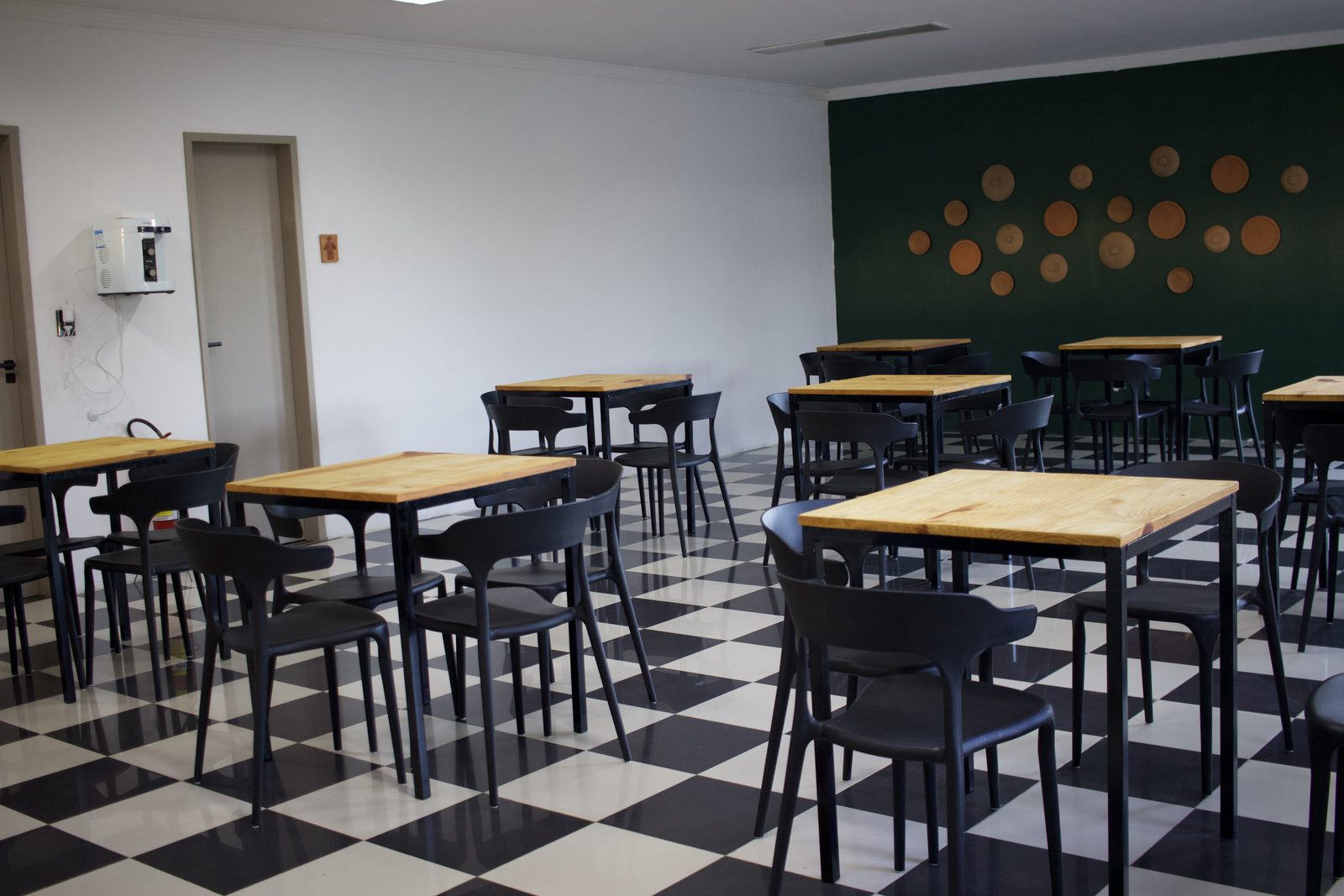 Salão do café da manhã com piso xadrez preto e branco, mesas de madeira e parede verde escuro