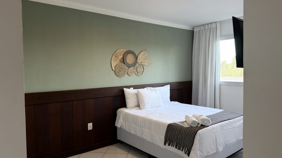 Vista lateral de um quarto com cama de casal, TV na parede e cortina fechando a janela