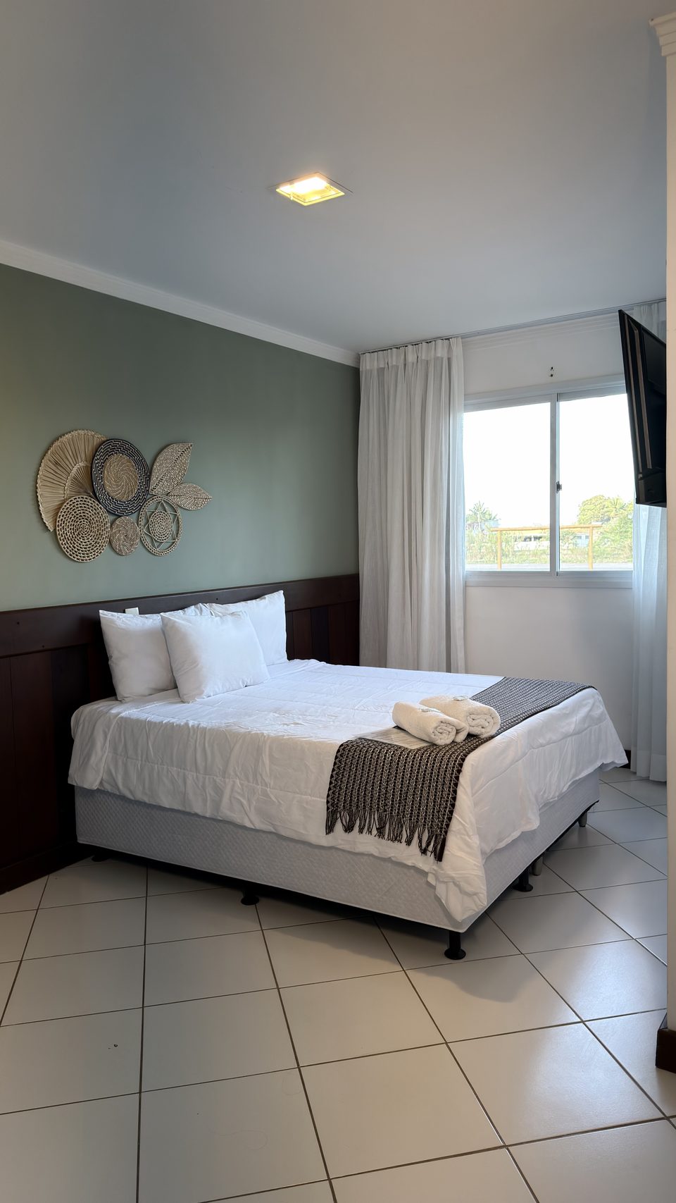 Quarto com cama de casal visto da porta: parede verde sálvia, cama arrumada e janela com cortina clara