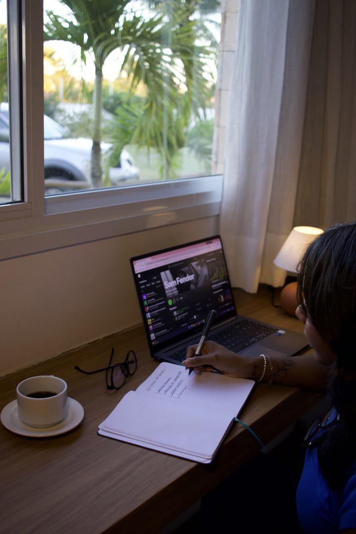 Hóspede trabalhando no notebook na escrivaninha do quarto, com café e caderno, ao lado da janela